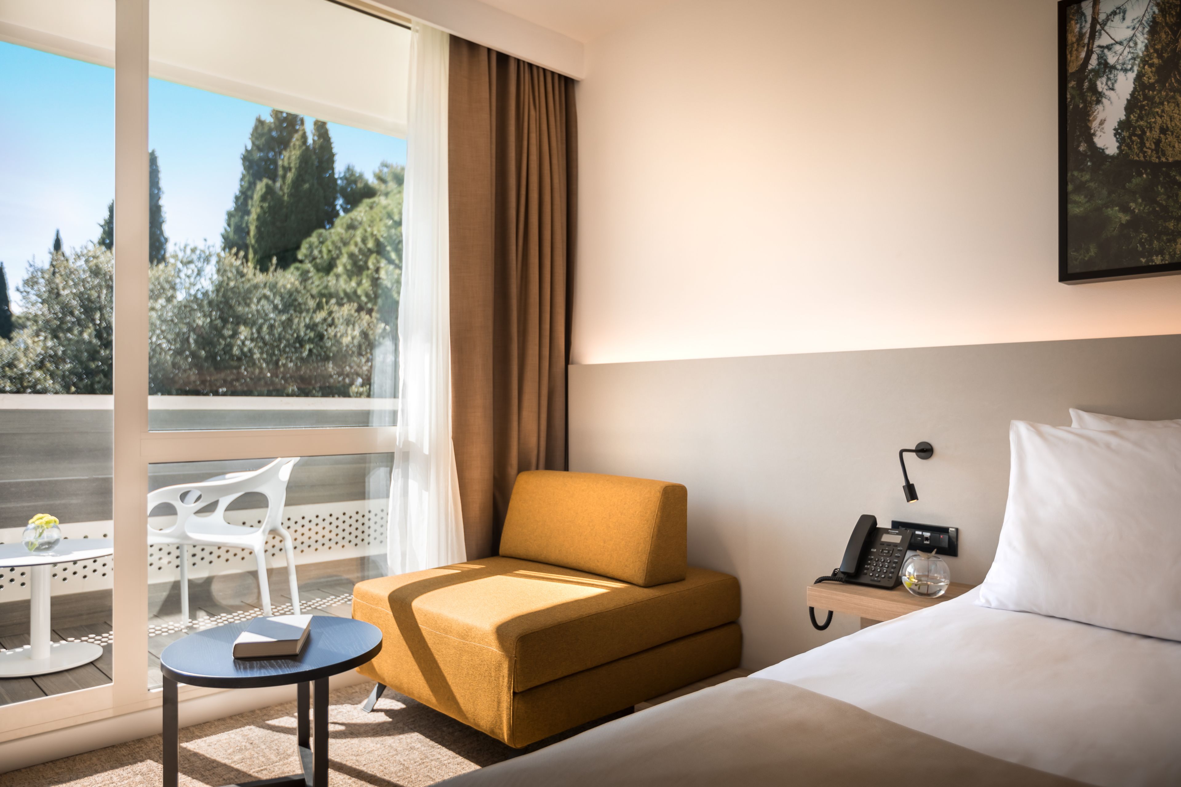 Habitación de hotel con cama, una mesita con un libro, una silla, una mesita de noche con un teléfono y una ventana con cortinas.