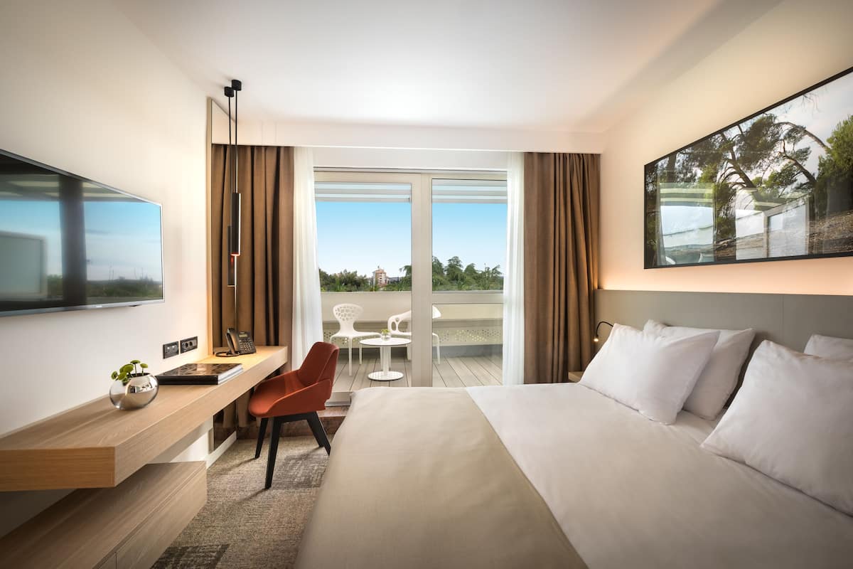 A modern hotel room with a large bed, a desk with a chair, a flat-screen TV, and a balcony with outdoor seating.