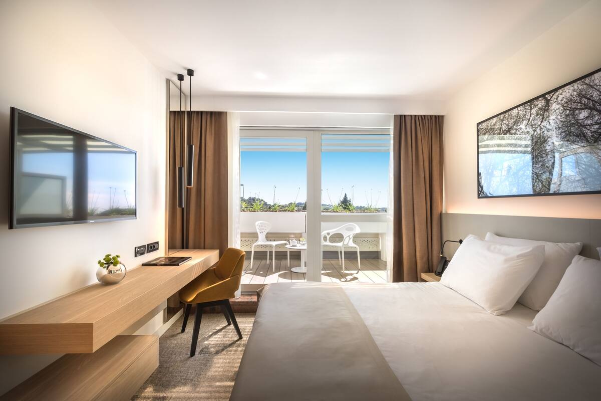 A modern hotel room with a large bed, a desk with a chair, a flat-screen TV, a large window with a view of the city, and a painting on the wall.
