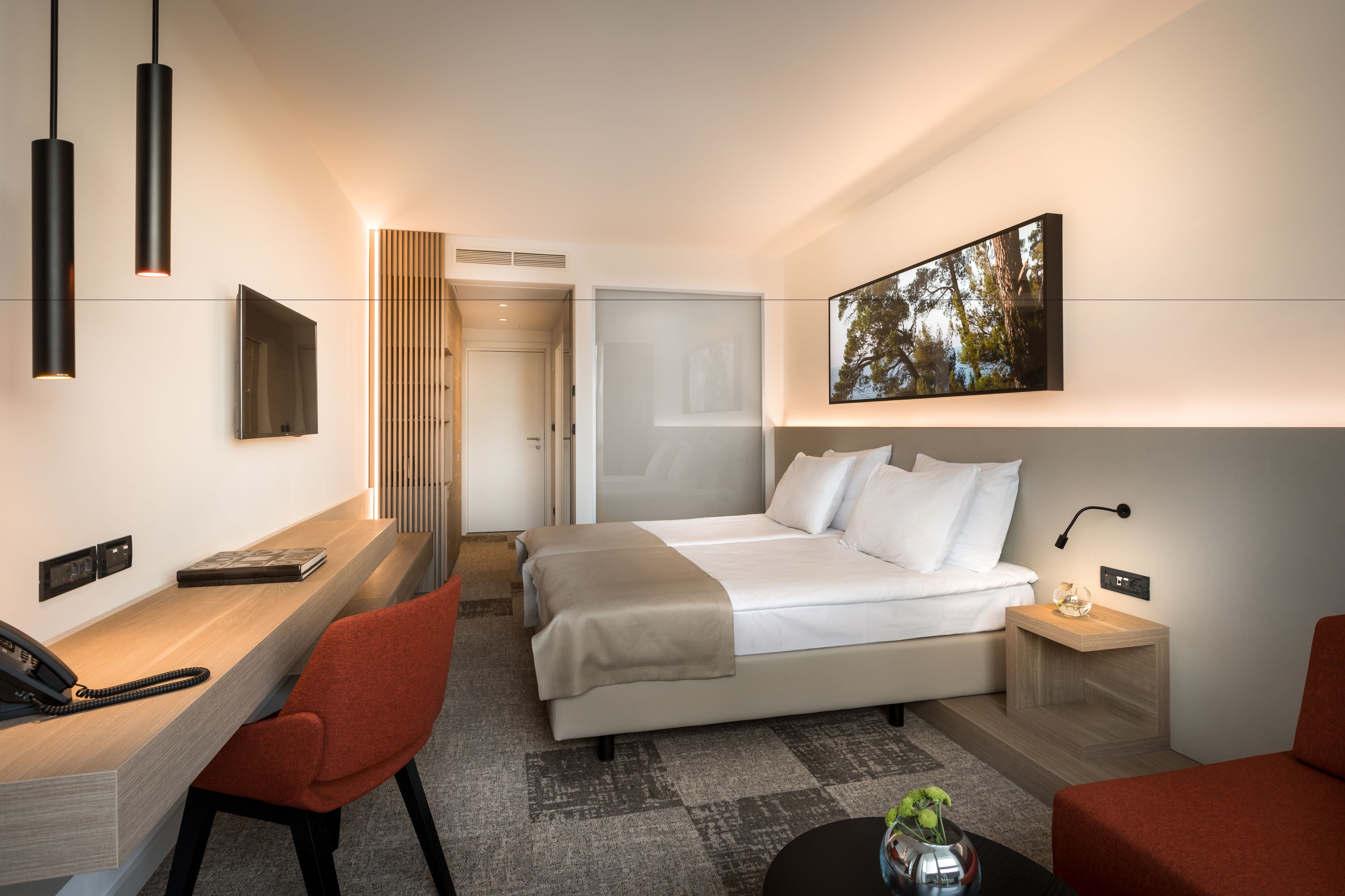 A modern hotel room with a bed, desk, and chair, a small table with a vase, and a painting on the wall.