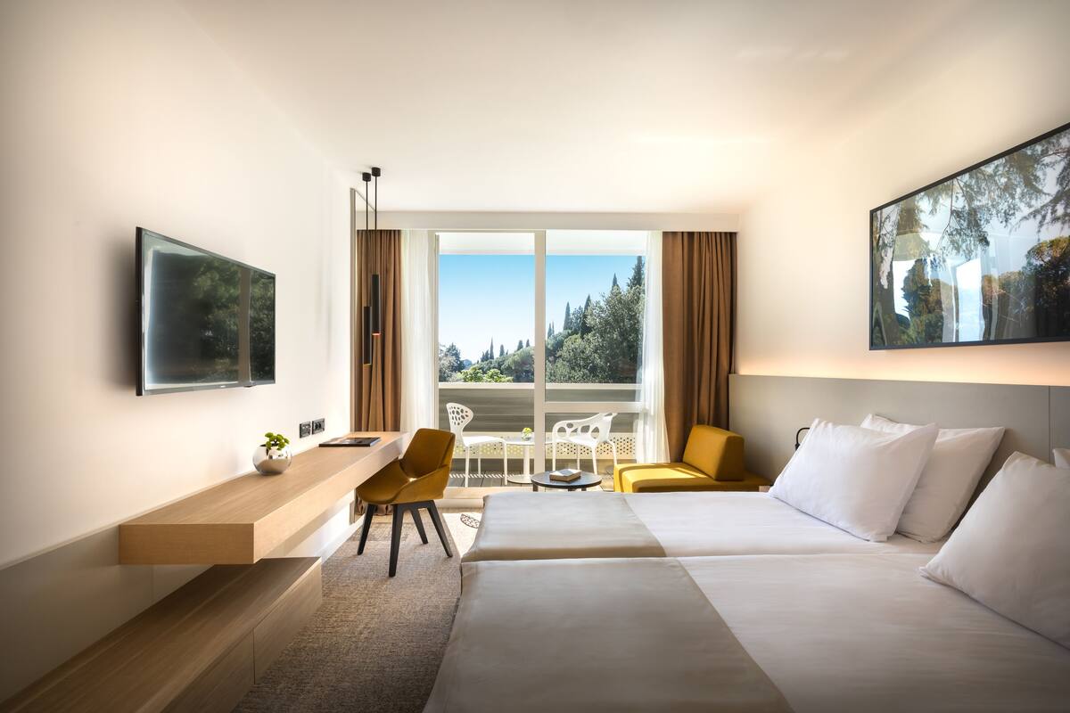A modern hotel room with a bed, a desk, a chair, and a balcony with a view of trees and buildings.