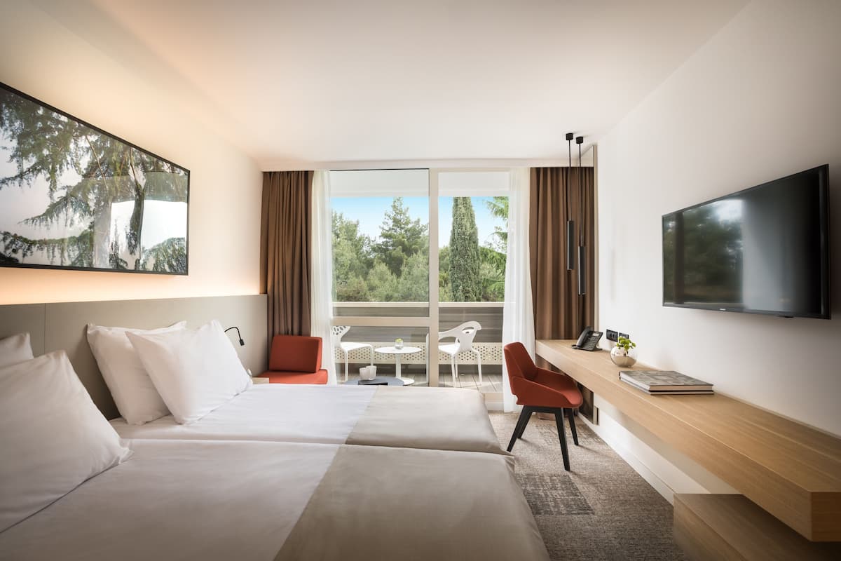 A hotel room with two beds, a desk with a chair, a TV, and a large window with curtains.