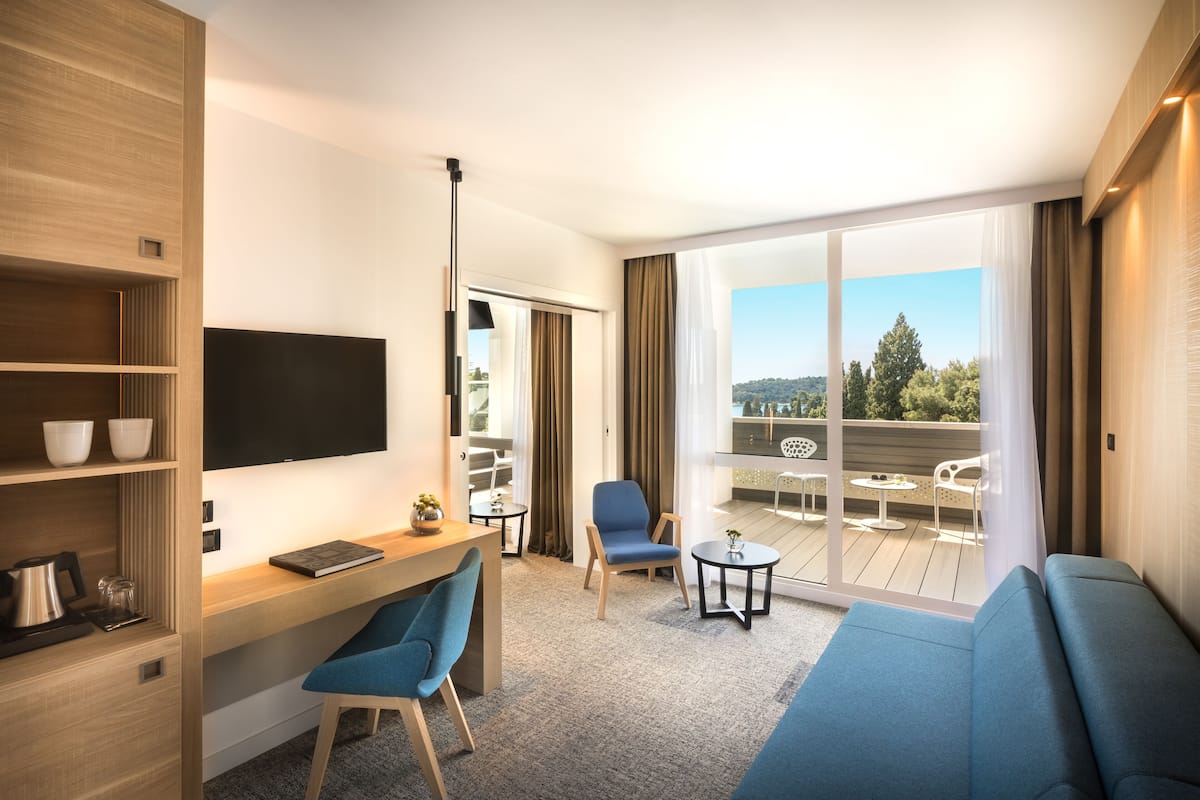 A modern hotel room with a flat-screen TV, a desk with a chair, a sofa, and a balcony with outdoor seating.