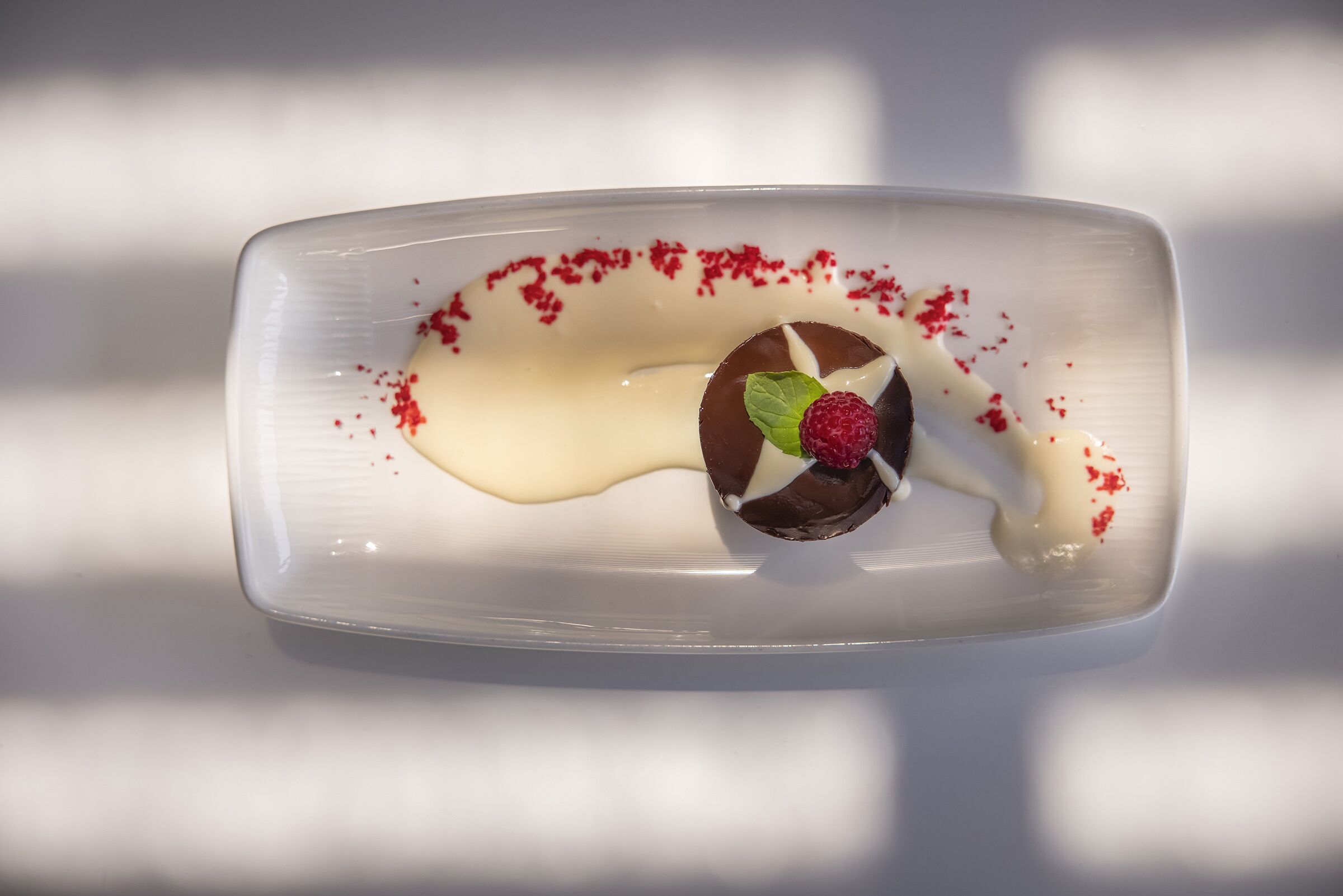 A dessert on a white rectangular plate with a chocolate sphere, a raspberry, and a mint leaf, surrounded by white sauce and red powder.