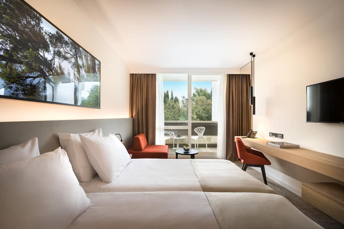 A hotel room with a large bed, a desk, a chair, a TV, and a balcony with a view of trees.
