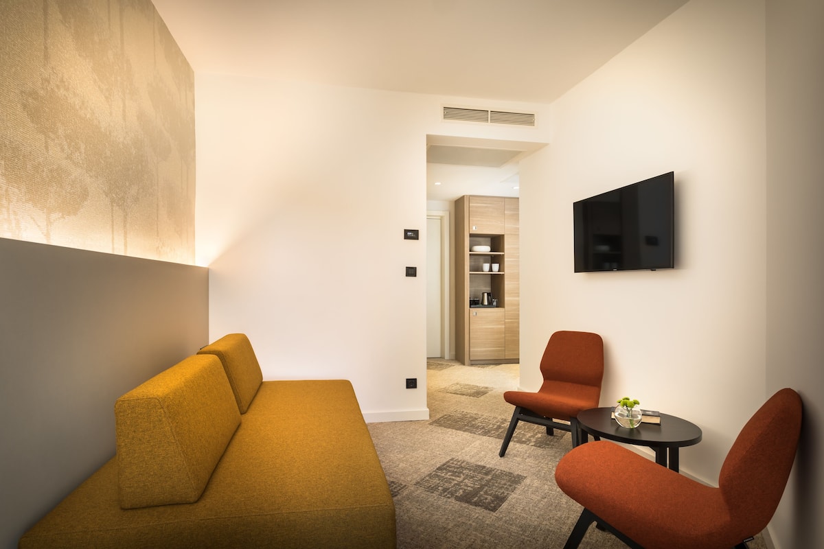 A modern living room with a sofa, armchairs, a small table, and a flat-screen TV mounted on the wall.