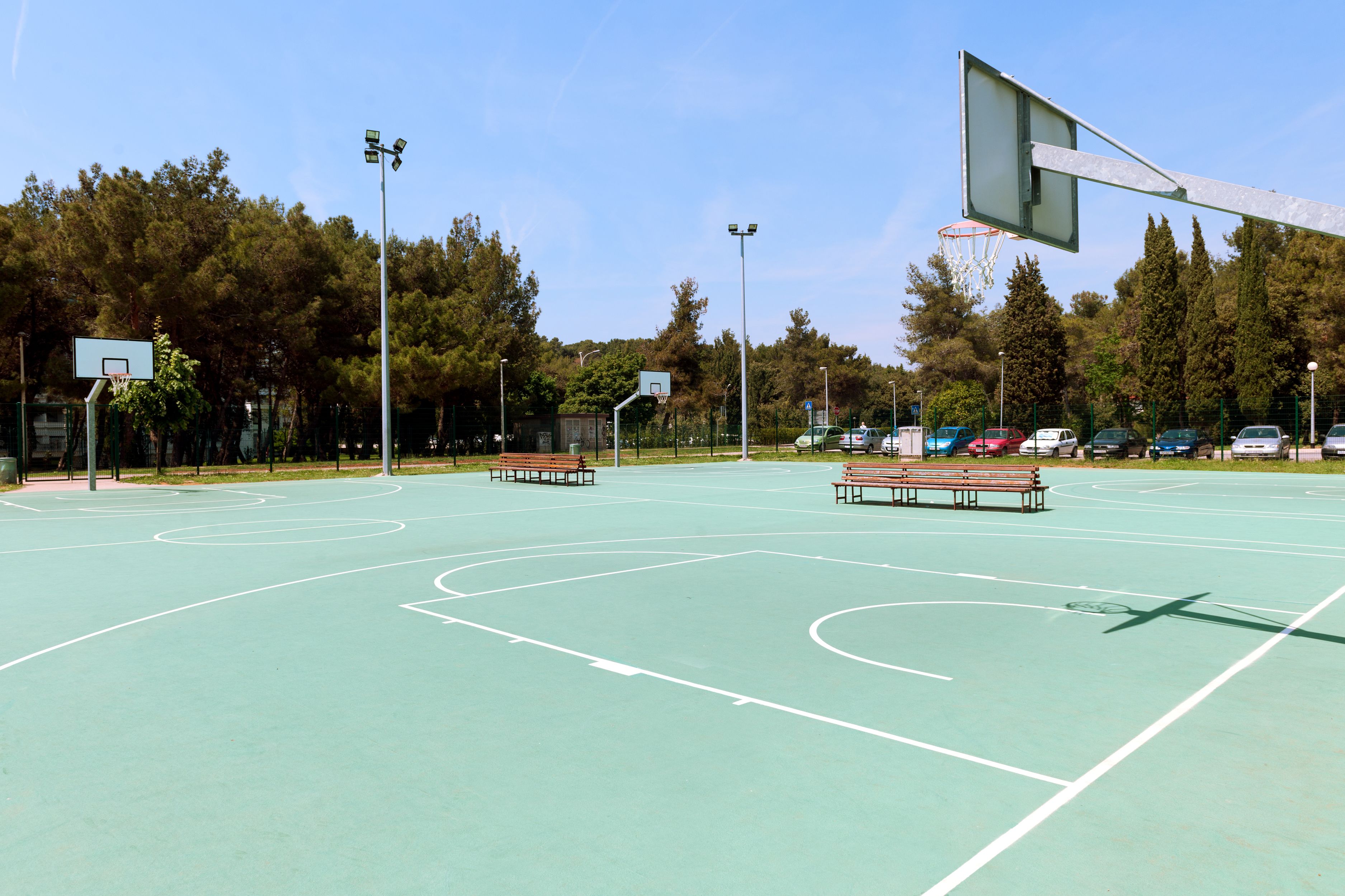 Una cancha de baloncesto al aire libre con varias bancas, aros de baloncesto y un estacionamiento con coches.