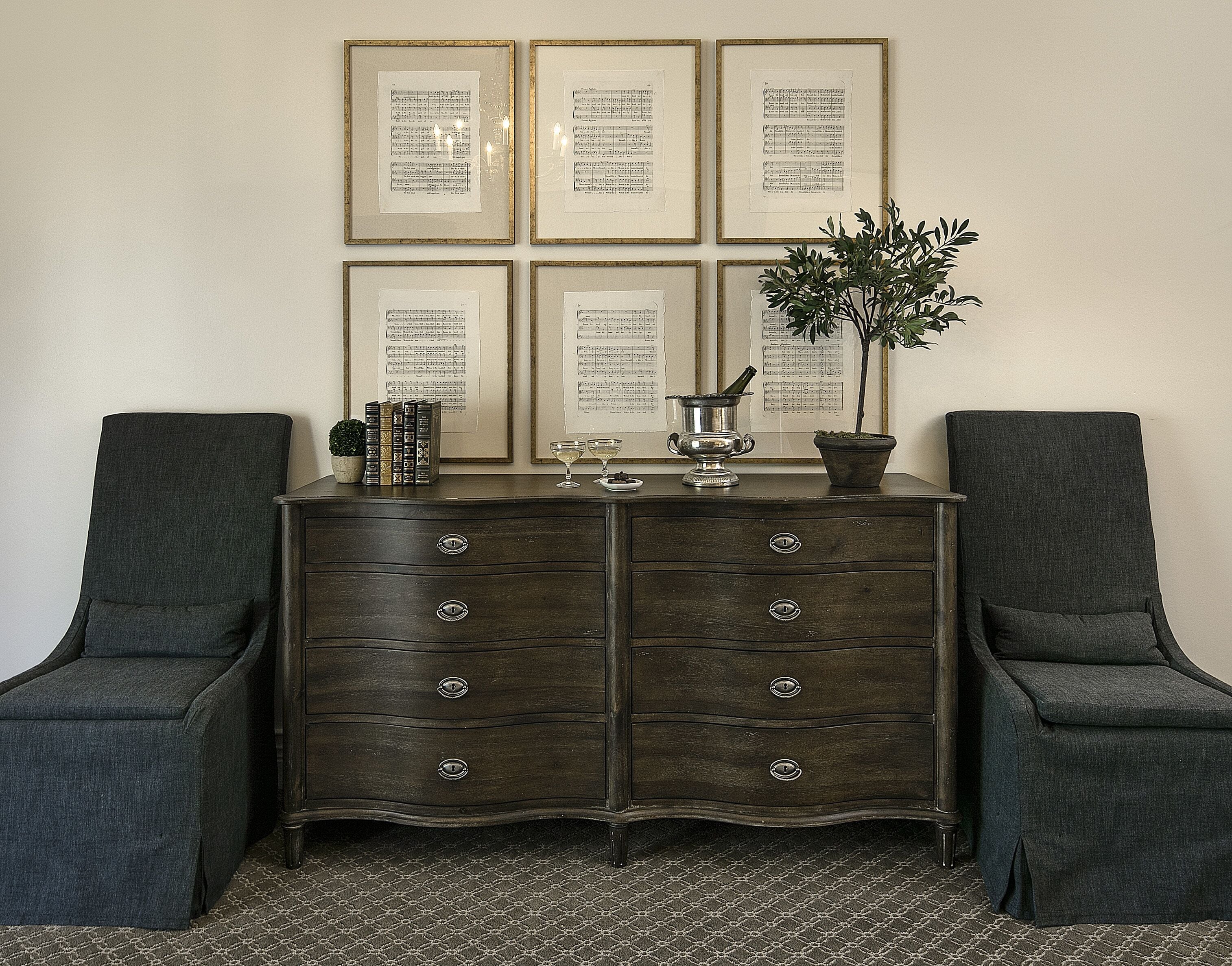 A dark wooden dresser with six drawers, flanked by two upholstered armchairs. Behind the dresser, there are six framed sheet music pieces on the wall.