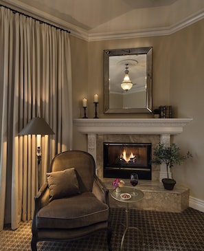 A cozy living room with a fireplace, a comfortable armchair, a small table with a glass of wine, and a potted plant.