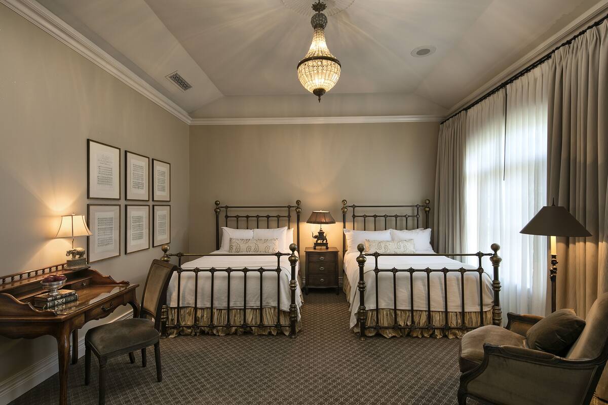 A hotel room with two beds, a desk, a chair, a lamp, and a chandelier.