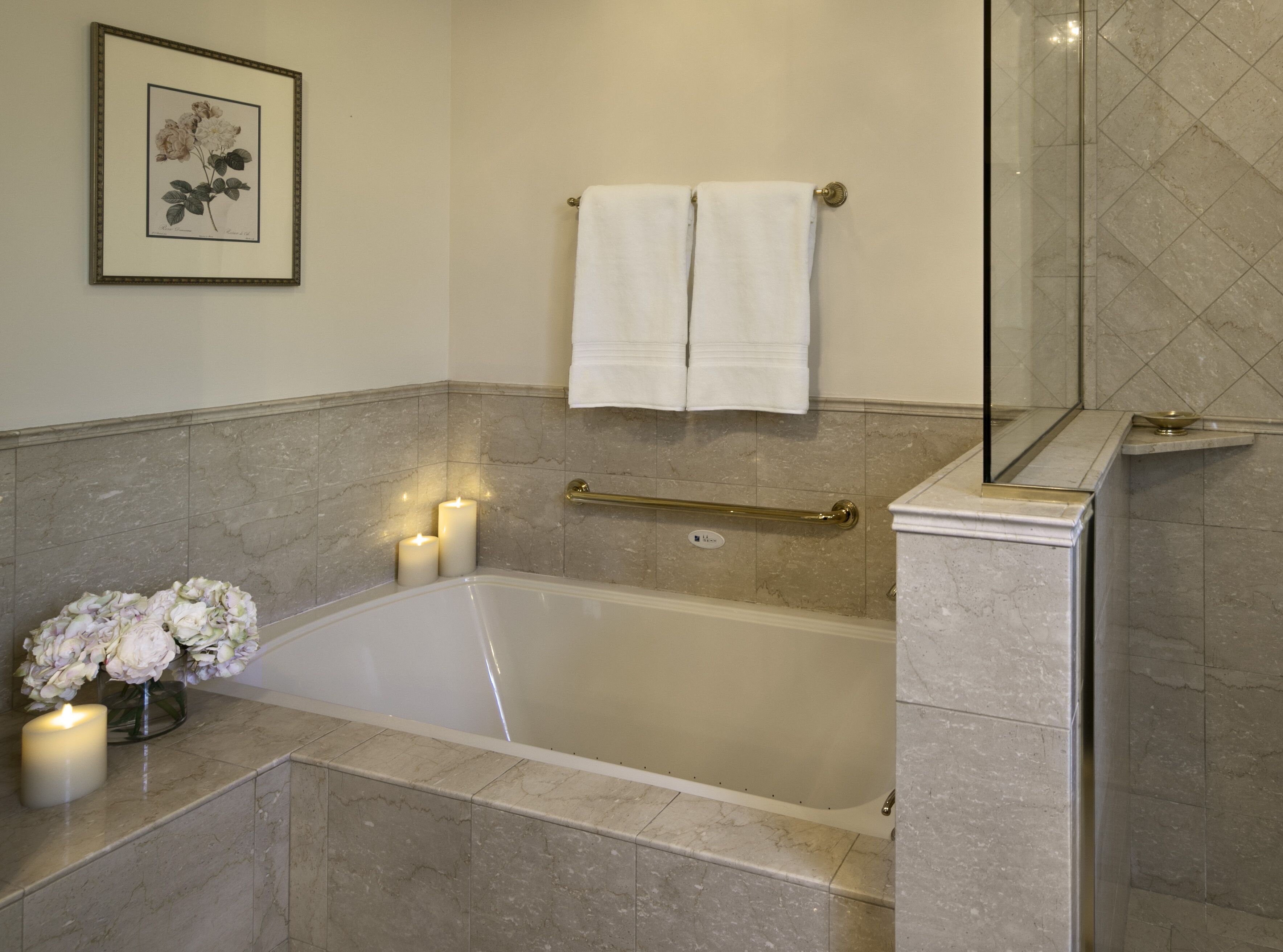 A bathroom with a bathtub, two lit candles, a framed floral artwork, and a towel rack.