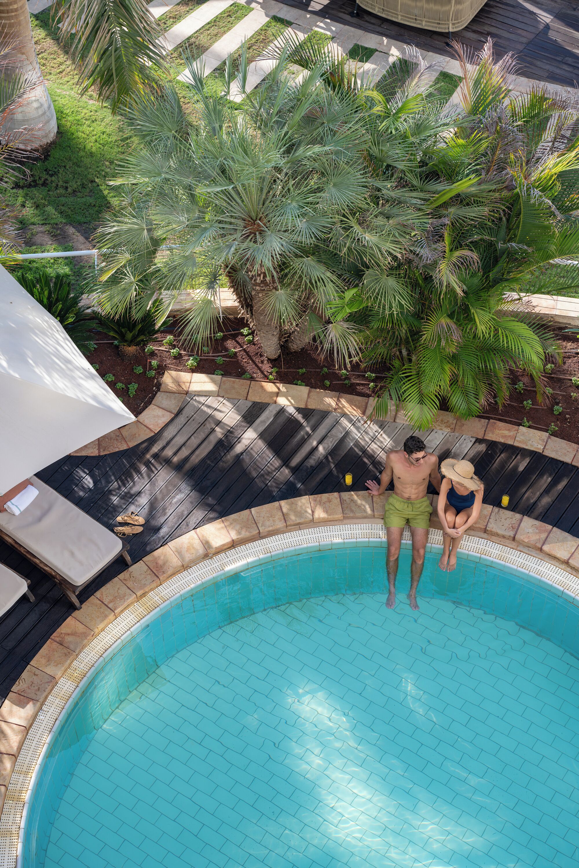 A swimming pool with a tiled edge, surrounded by a garden with palm trees and a white umbrella.