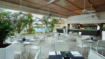 Un área de comedor al aire libre con sillas y mesas blancas, una piscina al fondo y una zona de bar donde un chef prepara comida.