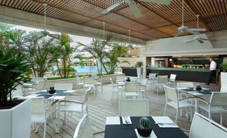 Un área de comedor al aire libre con sillas y mesas blancas, una piscina al fondo y una zona de bar donde un chef prepara comida.
