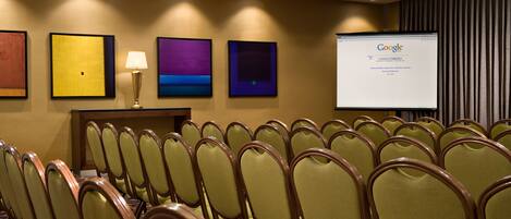 Un salón de conferencias con filas de sillas, una pantalla de proyector mostrando Google y obras de arte enmarcadas en la pared.