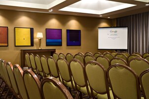 Un salón de conferencias con filas de sillas, una pantalla de proyector mostrando Google y obras de arte enmarcadas en la pared.