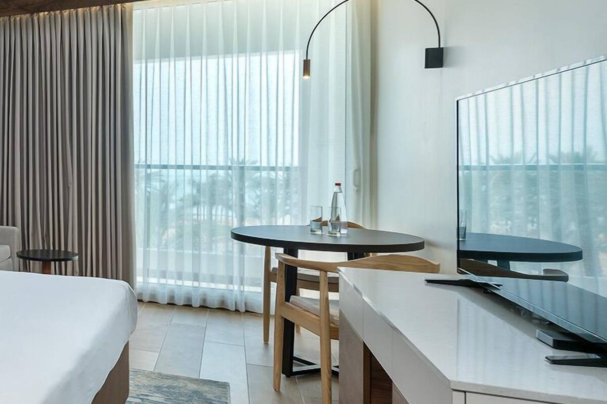 Ett hotellrum med en säng, ett skrivbord med en stol, en TV och utsikt över palmerna genom stora fönster.