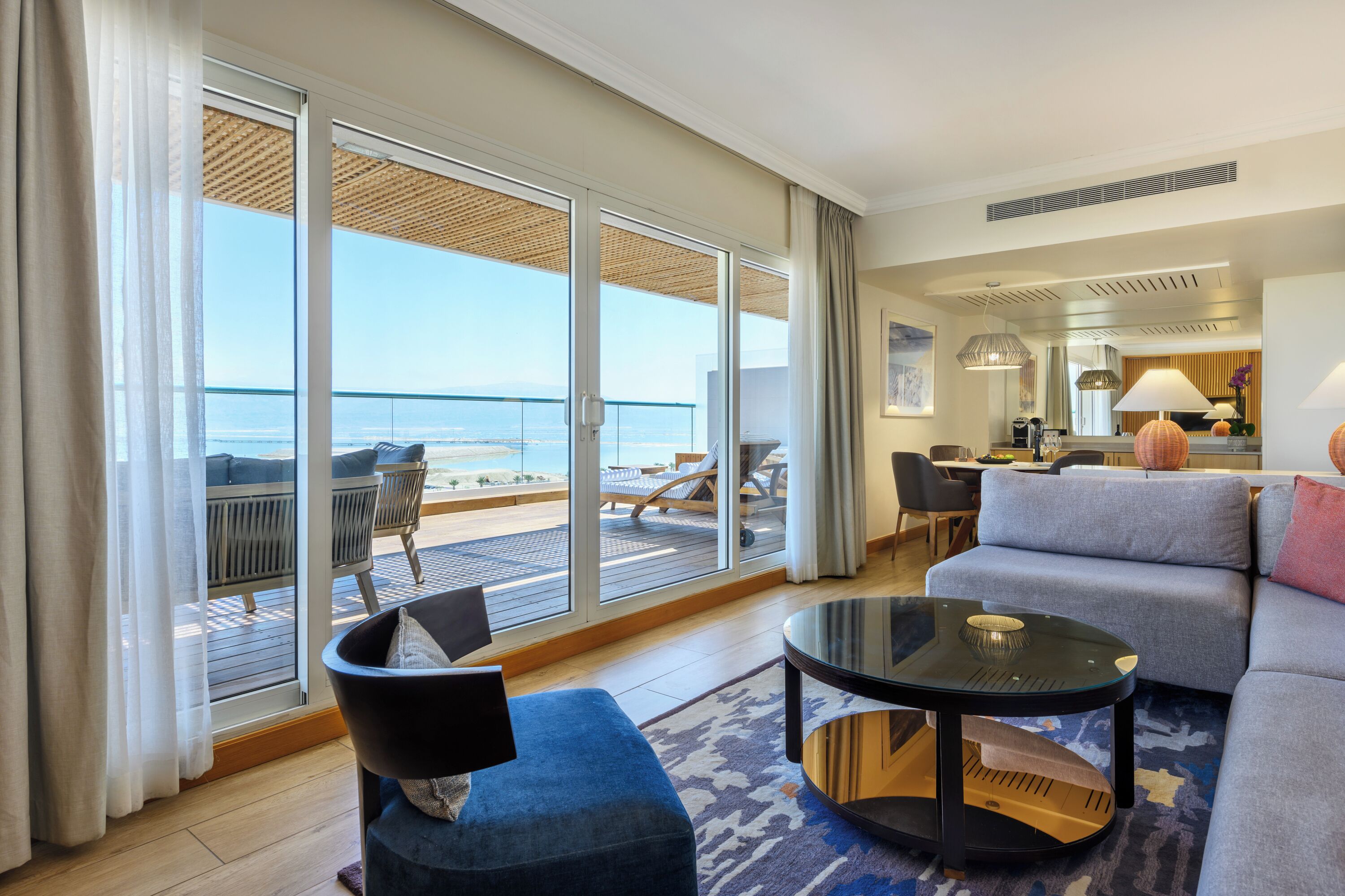 A modern hotel room with a balcony overlooking the sea, a sofa, a coffee table, and a dining area.