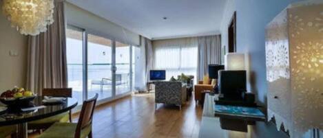 A hotel room with a dining area, a TV, and a large window with a view of the sea.