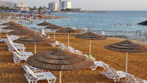 Une plage bordée de rangées de chaises longues blanches et d’ombrelles en chaume, un espace piscine et, au loin, une vue sur la ville.