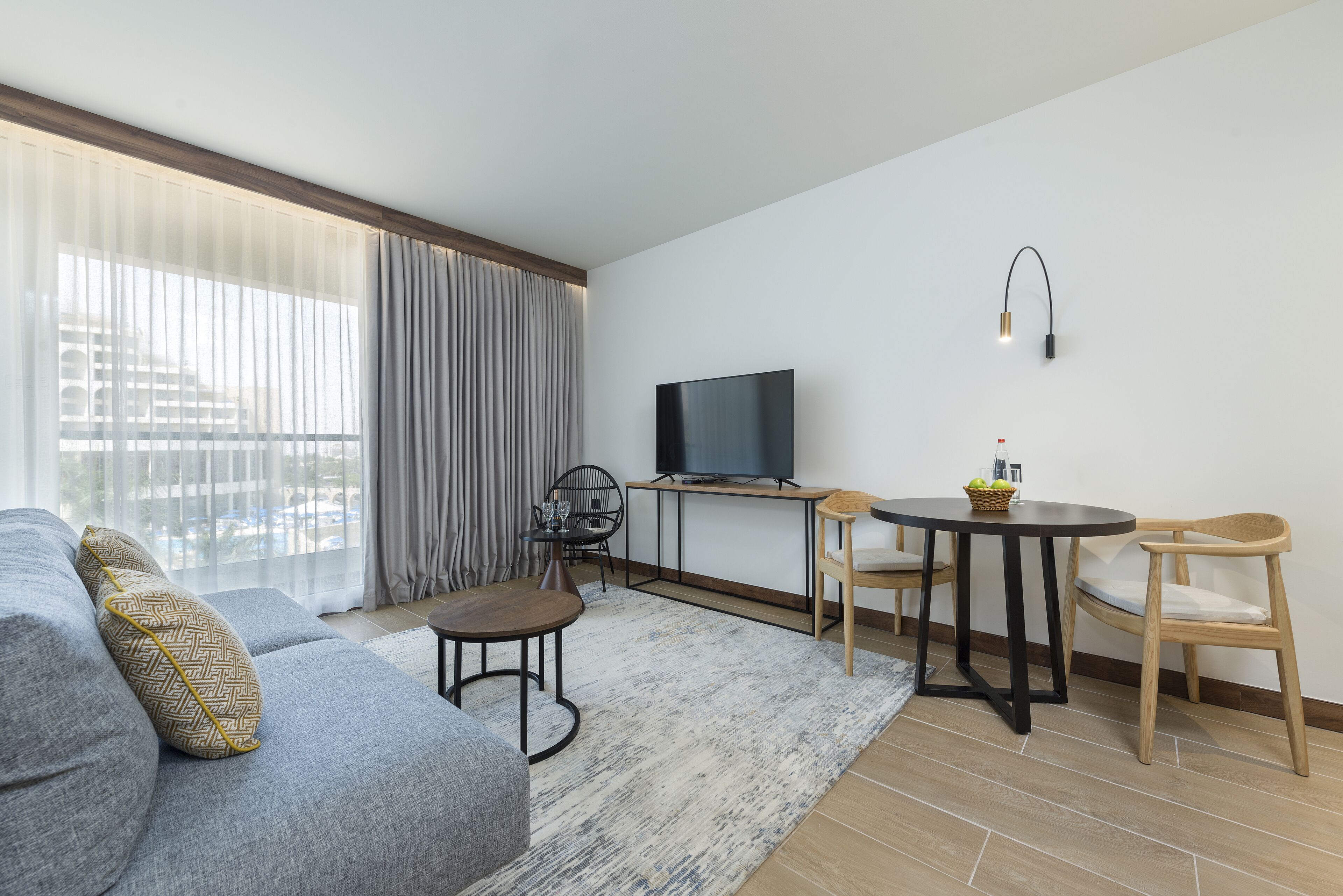 A modern living room with a grey sofa, a small round table, a TV on a stand, and a window with curtains.