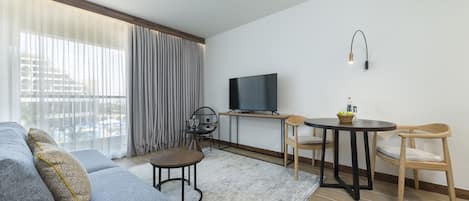 A modern living room with a grey sofa, a small round table, a TV on a stand, and a window with curtains.
