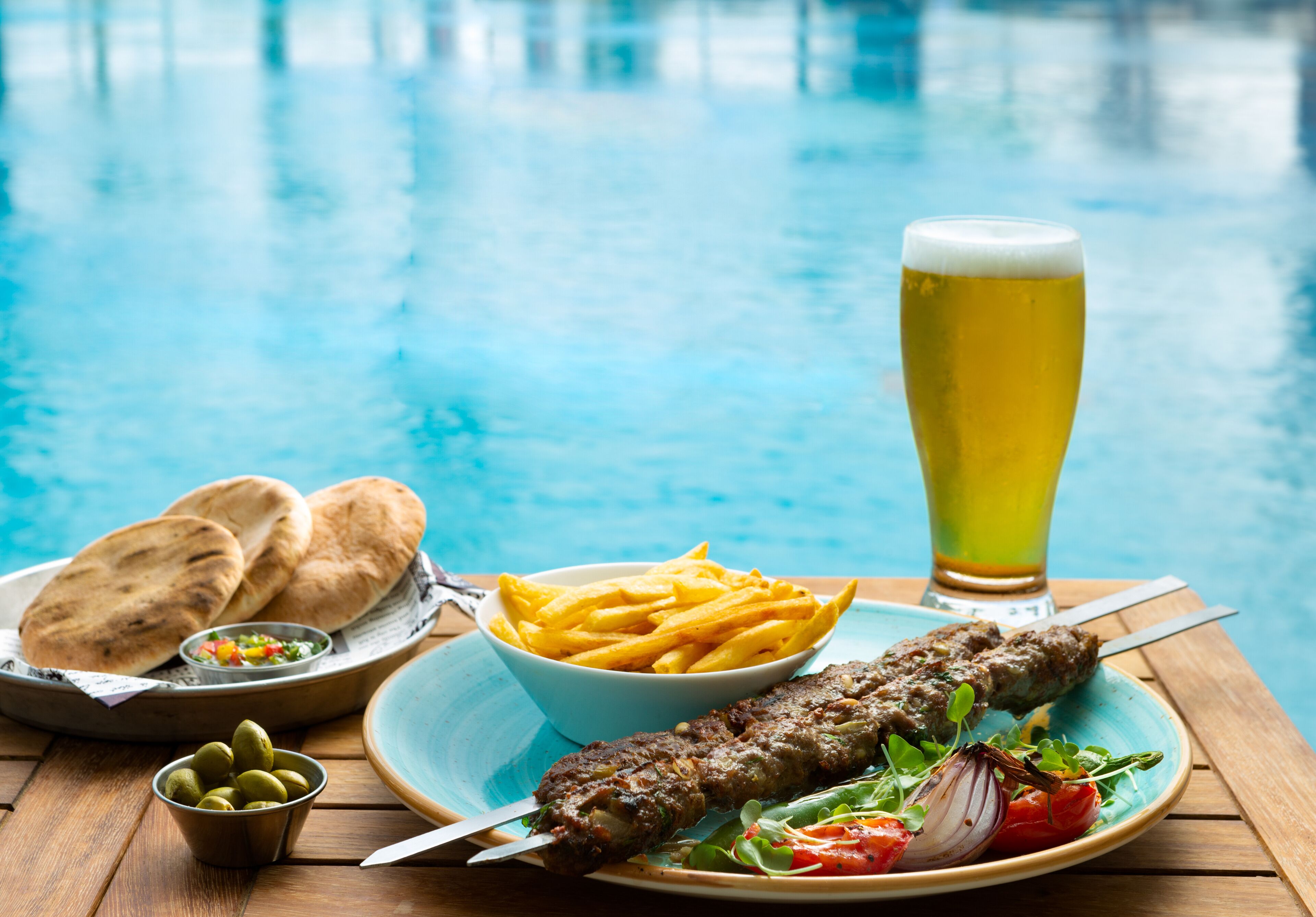 A meal with kebabs, fries, flatbread, and a glass of beer by a pool.