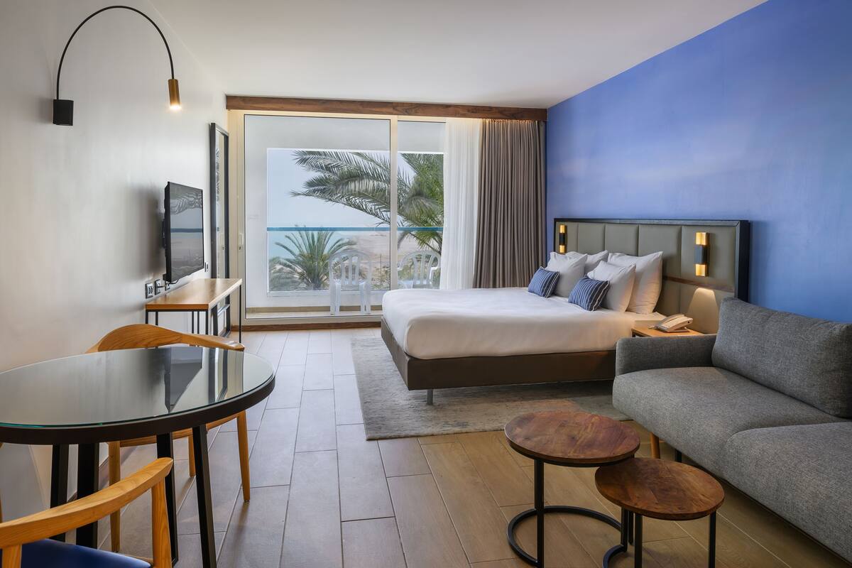 Ett hotellrum med en säng, en soffa, en tv, ett bord och utsikt över stranden.