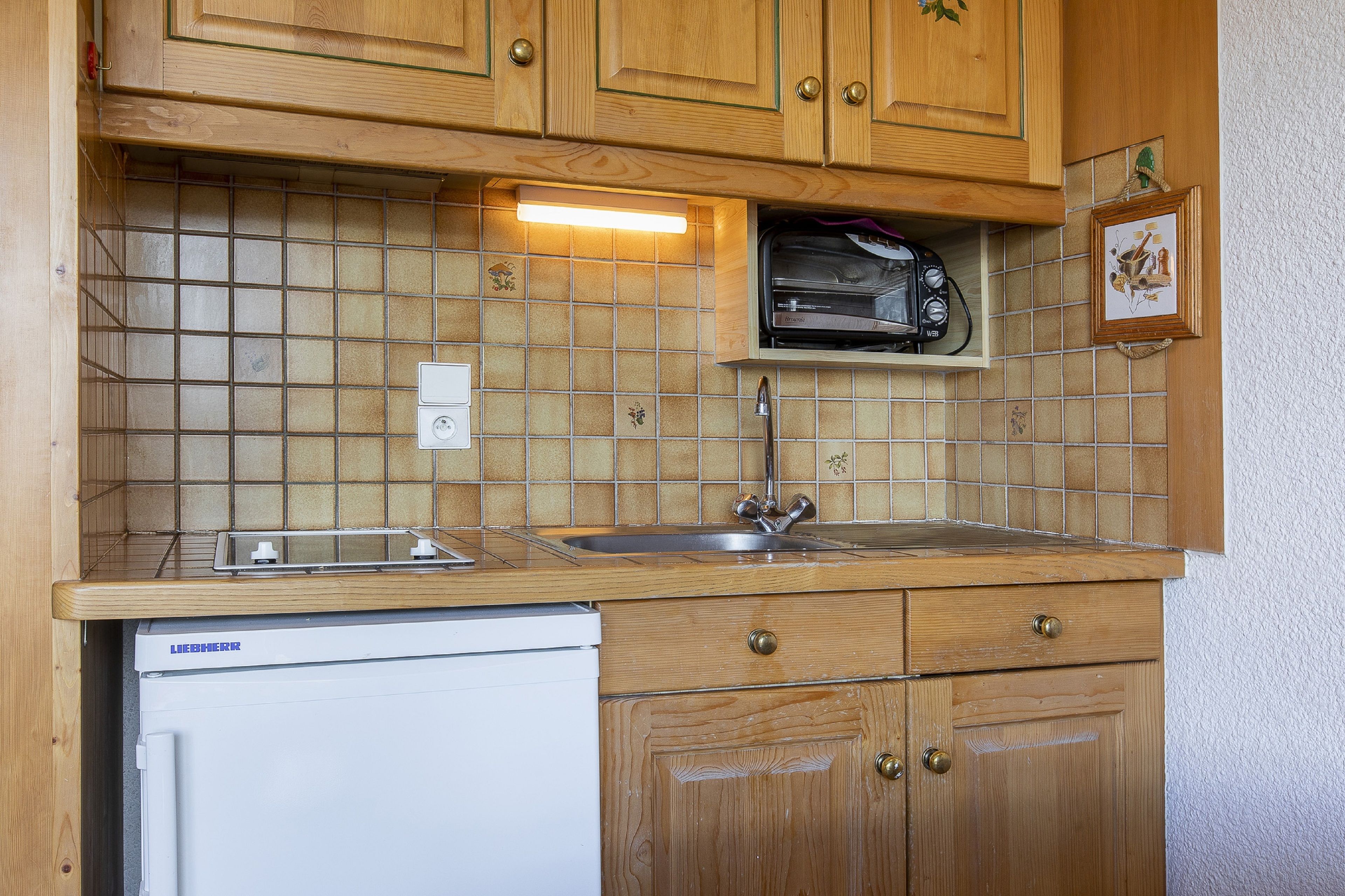 A compact kitchen with wooden cabinets, a built-in oven, a dishwasher, and a tiled backsplash.