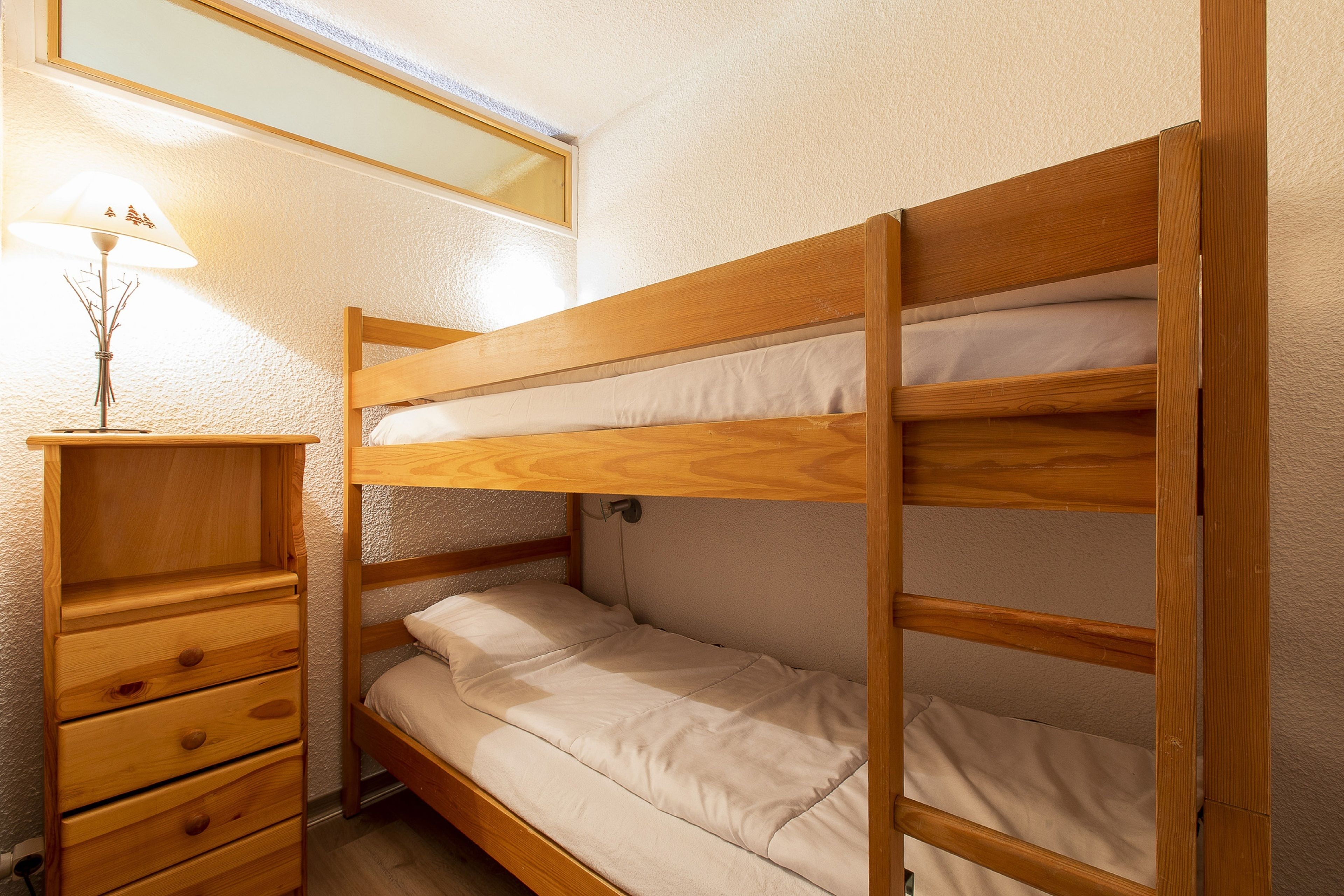 Een stapelbed met houten frame en witte beddengoed, een houten dressoir met lades, en een kleine tafel met een lamp.