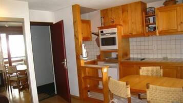 Een kleine keuken met houten kastjes, een inbouwoven en een eethoek met een houten tafel en stoelen.