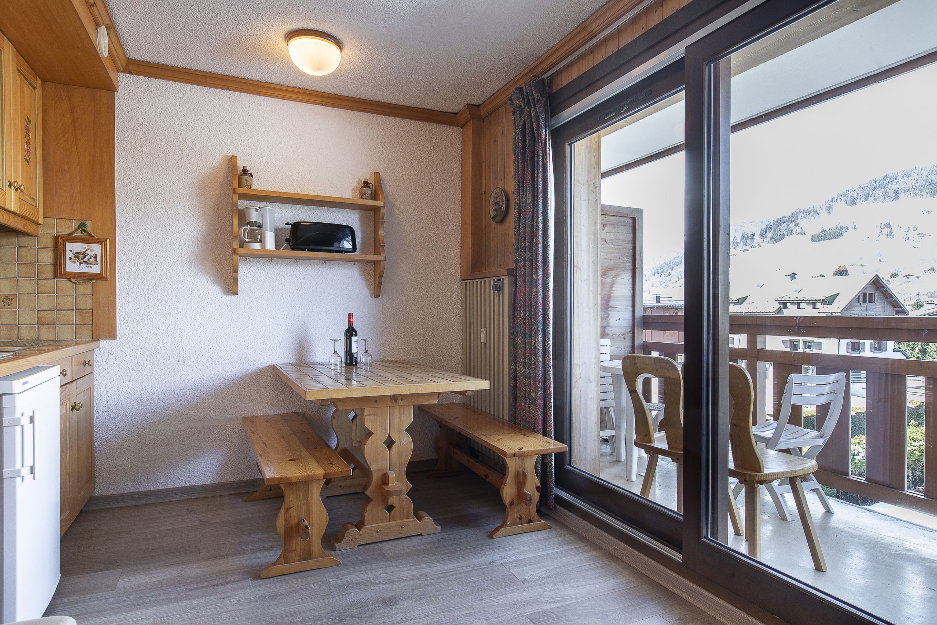 A wooden dining table with benches, a kitchen area with wooden cabinets, and a view of a snowy mountain through large windows.