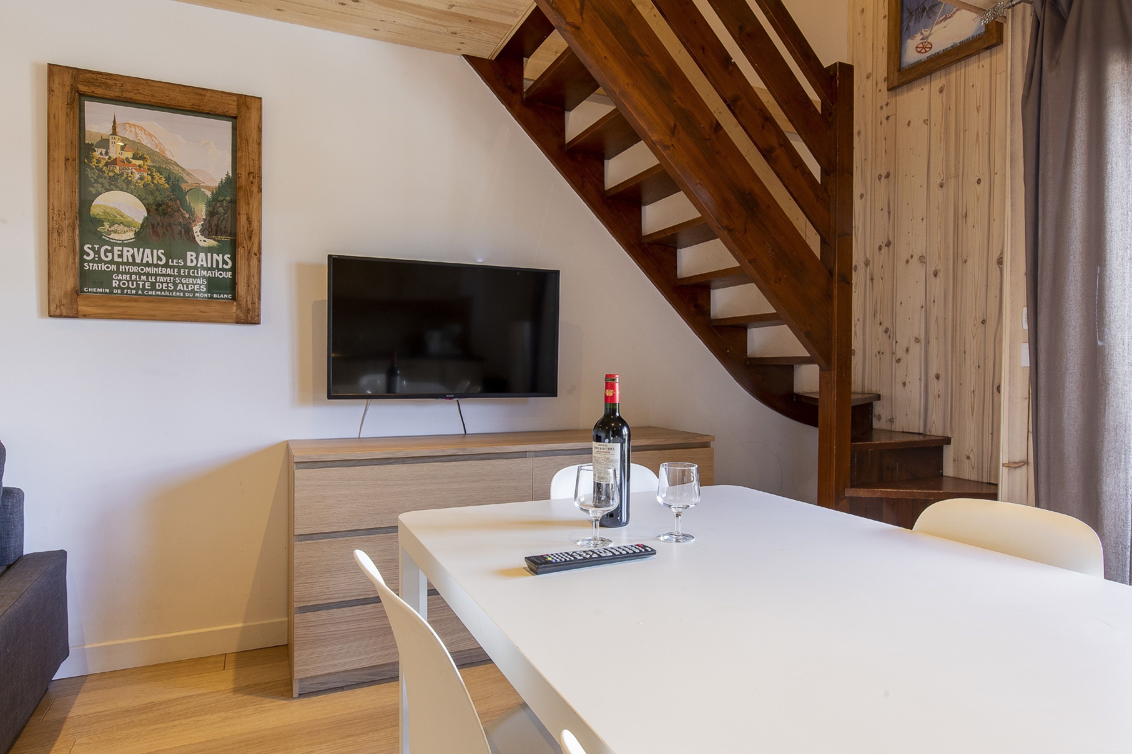 A dining area with a white table, chairs, a bottle of wine, and a remote control. A TV is mounted on the wall above the table.