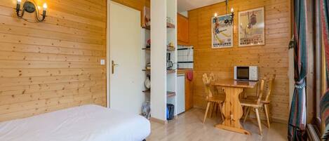 Habitación con paredes de madera, una cama, un pequeño rincón de cocina, una mesa de comedor con sillas y un televisor.