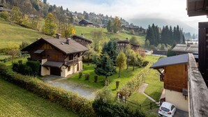 A traditional chalet-style building with a sloped roof, surrounded by a well-maintained garden and a view of a mountainous village. - Résidence Hôtel Rent (Megeve)