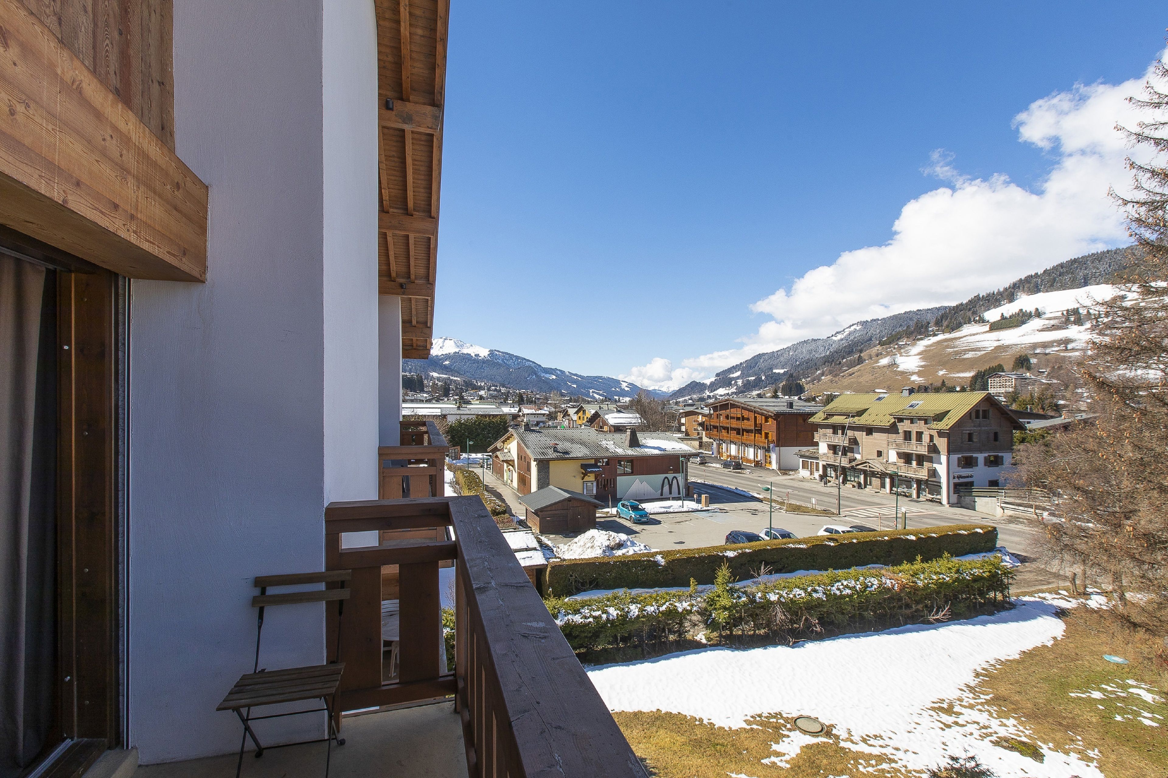 A balcony with a wooden railing overlooking a town with buildings and snow-covered mountains in the background.