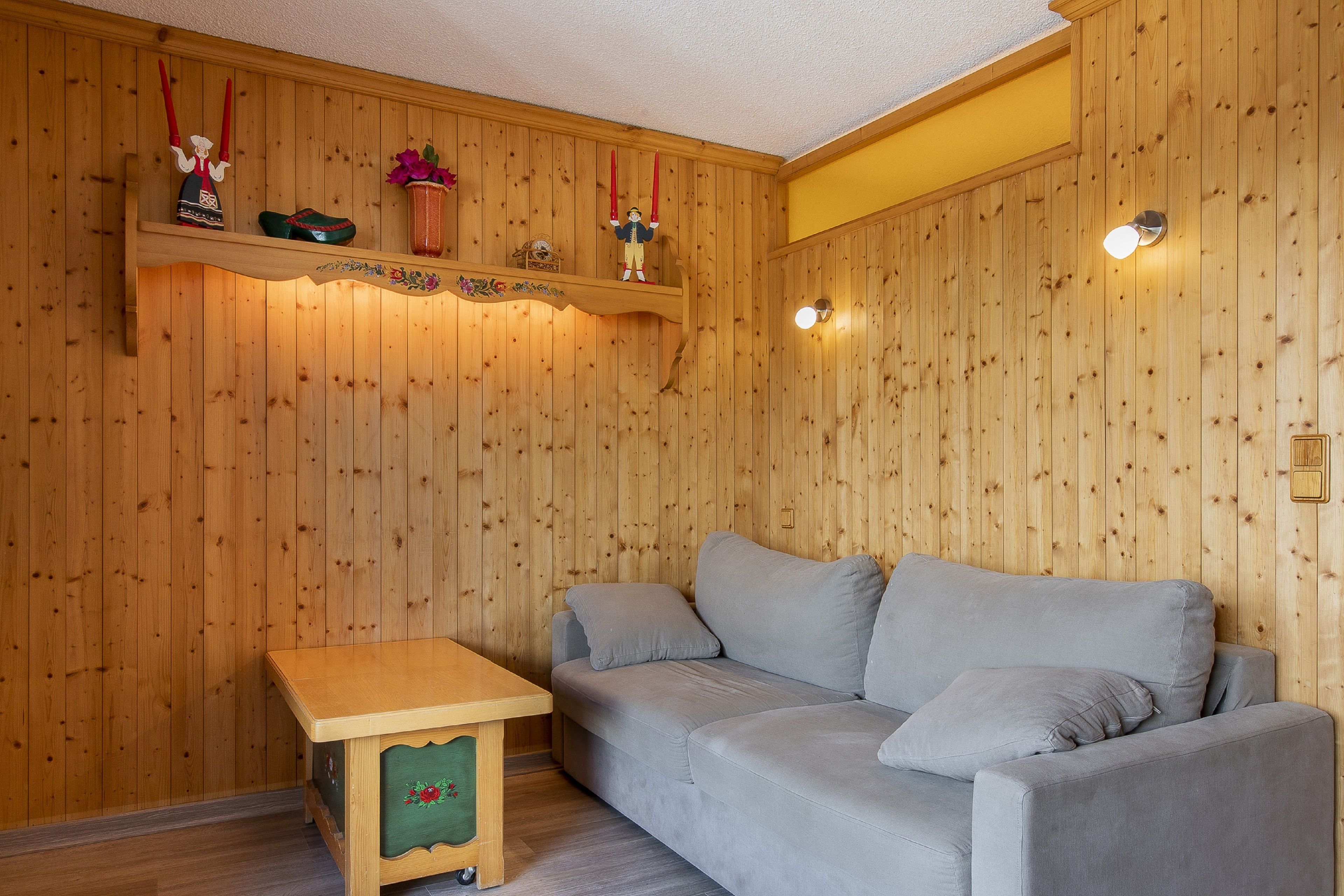 A cozy room with wooden panel walls, a grey sofa, a wooden table, and decorative items on shelves.