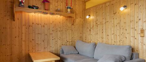 A cozy room with wooden panel walls, a grey sofa, a wooden table, and decorative items on shelves.