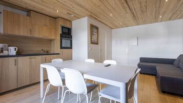 A modern kitchen with wooden cabinets, a white dining table with chairs, and a grey sofa in a living area.