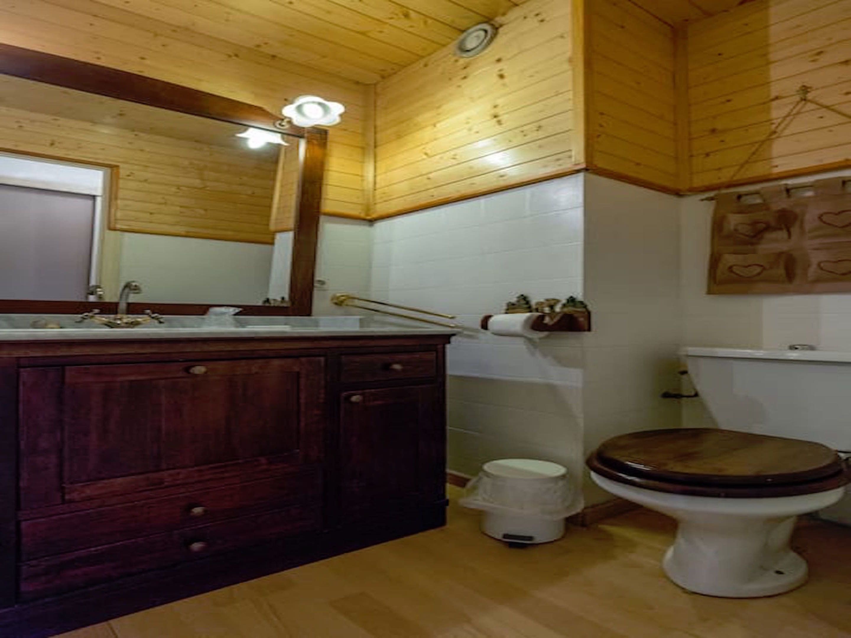 Un baño con paneles de madera, un tocador de madera con espejo, un inodoro y un cubo de basura.