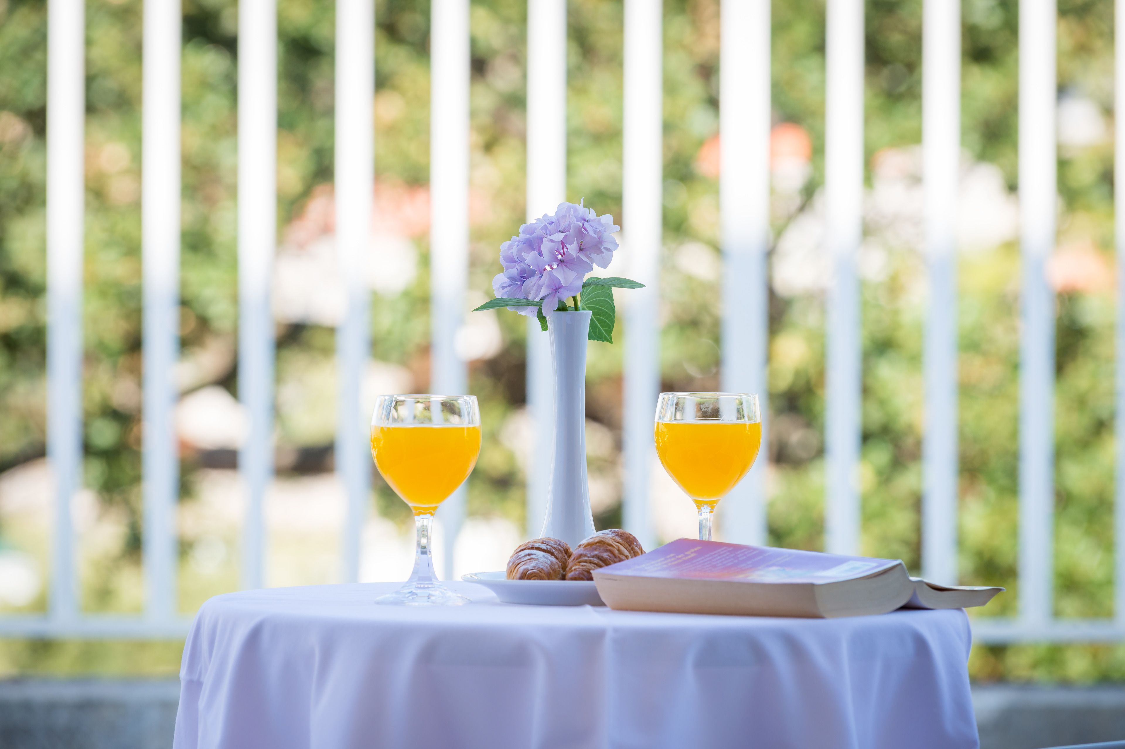 Una mesa con dos vasos de jugo de naranja, un libro y croissants sobre un mantel blanco.