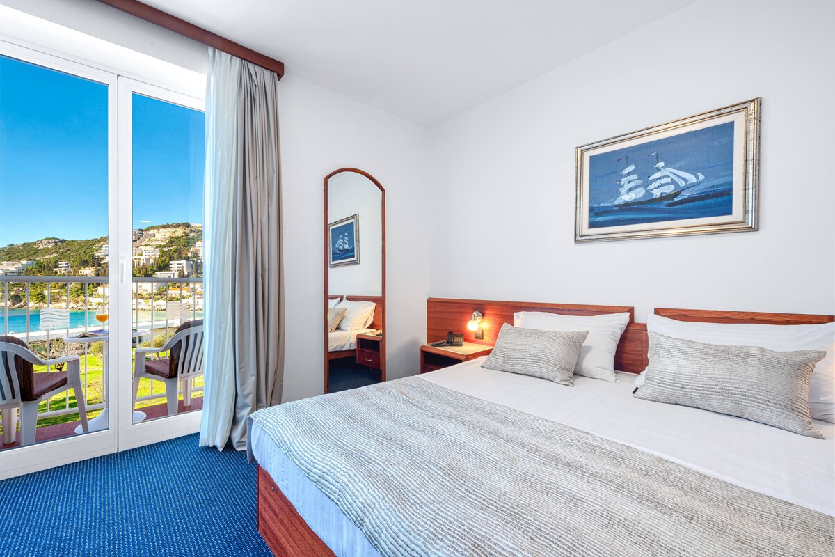 A hotel room with a large bed, a nightstand, a mirror, and a view of a pool and buildings.