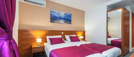 Een hotelkamer met twee bedden, een nachtkastje met een lamp, een airconditioningapparaat, een schilderij van een zeegezicht en een spiegel.
