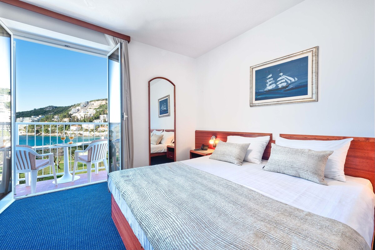 A hotel room with a large bed, a nightstand, a mirror, and a view of a coastal area.