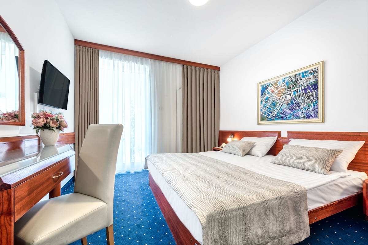 Quarto de hotel com cama, escrivaninha, cadeira, televisão e um quadro na parede.