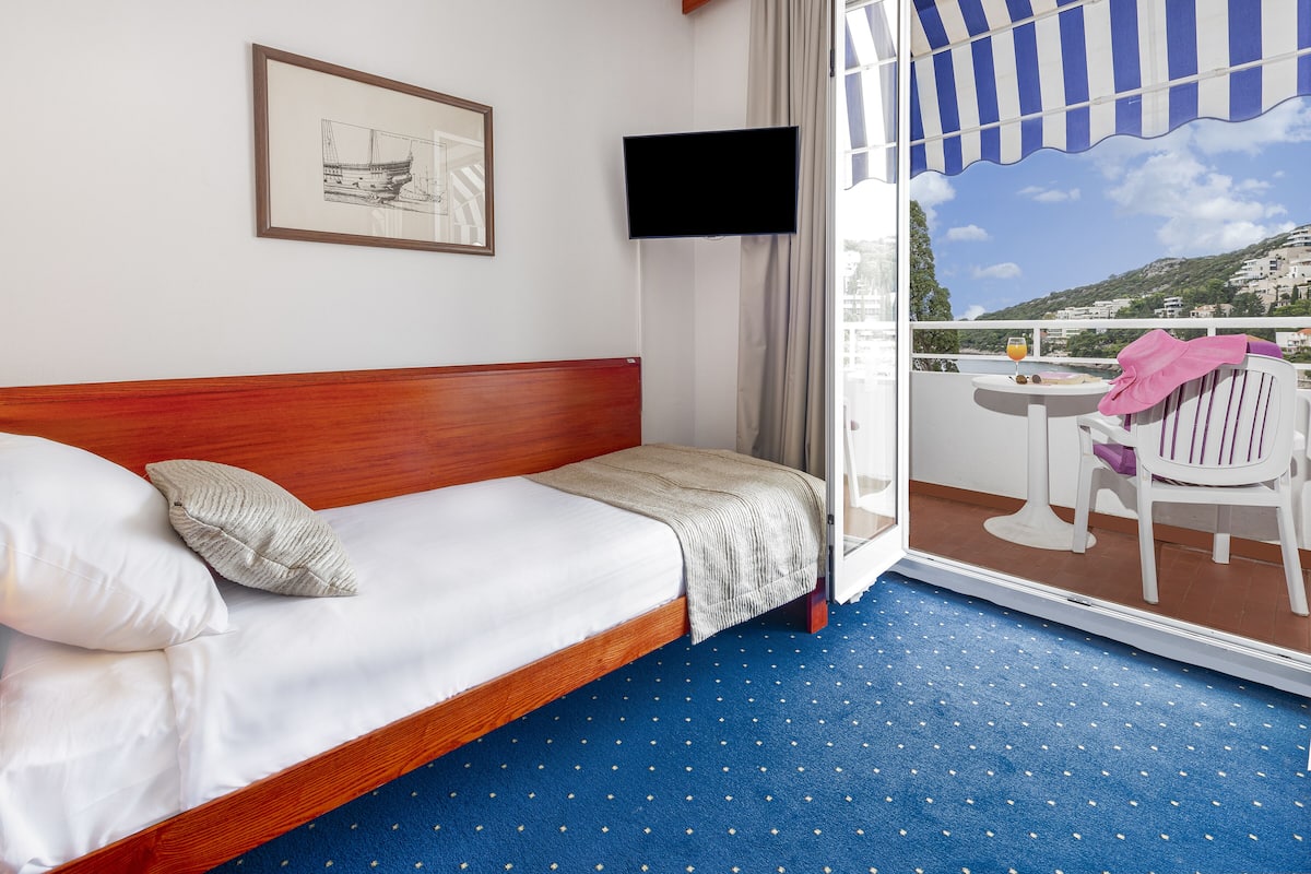 Quarto de hotel com cama, televisão na parede, varanda com mesa e cadeiras e vista para o mar.