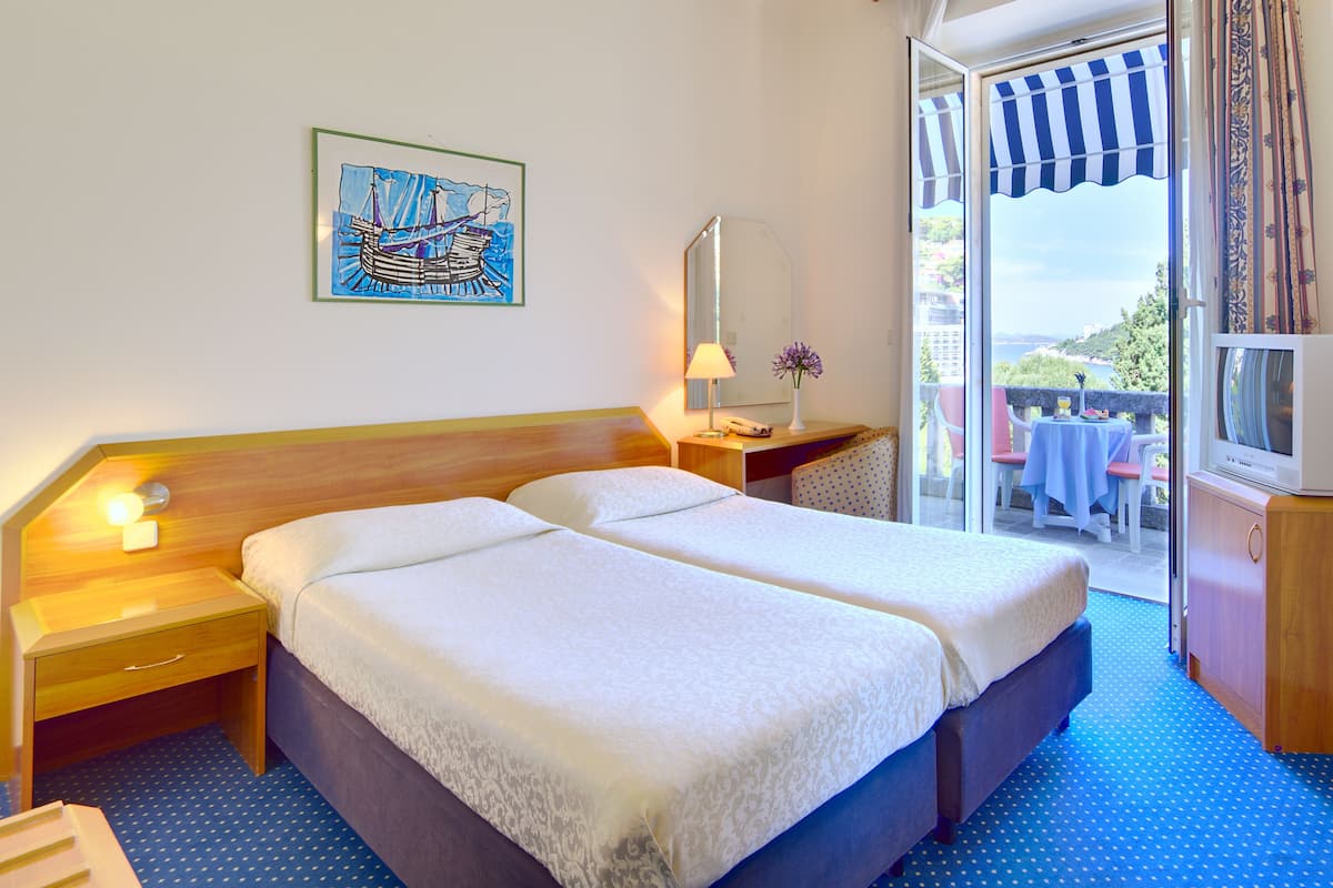 Quarto de hotel com duas camas, mesa de cabeceira, luminária, espelho e vista para a varanda externa.