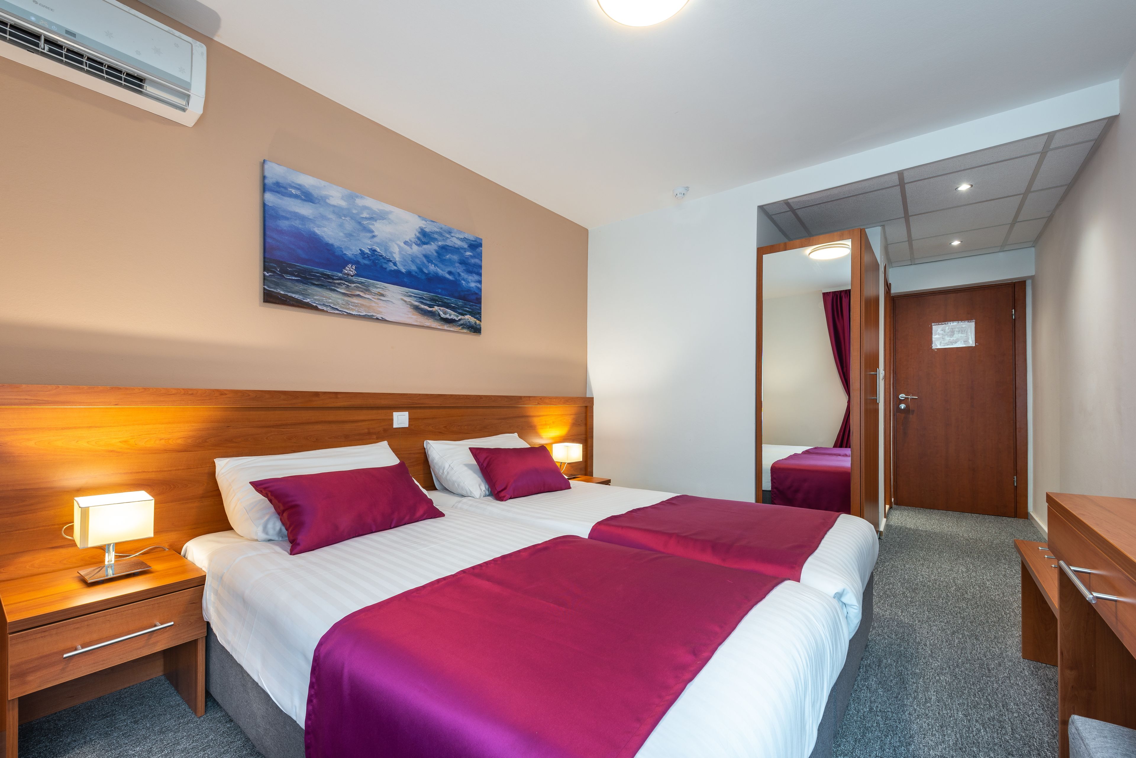 Quarto de hotel com uma cama grande, duas mesas de cabeceira, uma mesinha de apoio, um painel de cama de madeira, um quadro com vista para o mar, uma porta e um espelho.