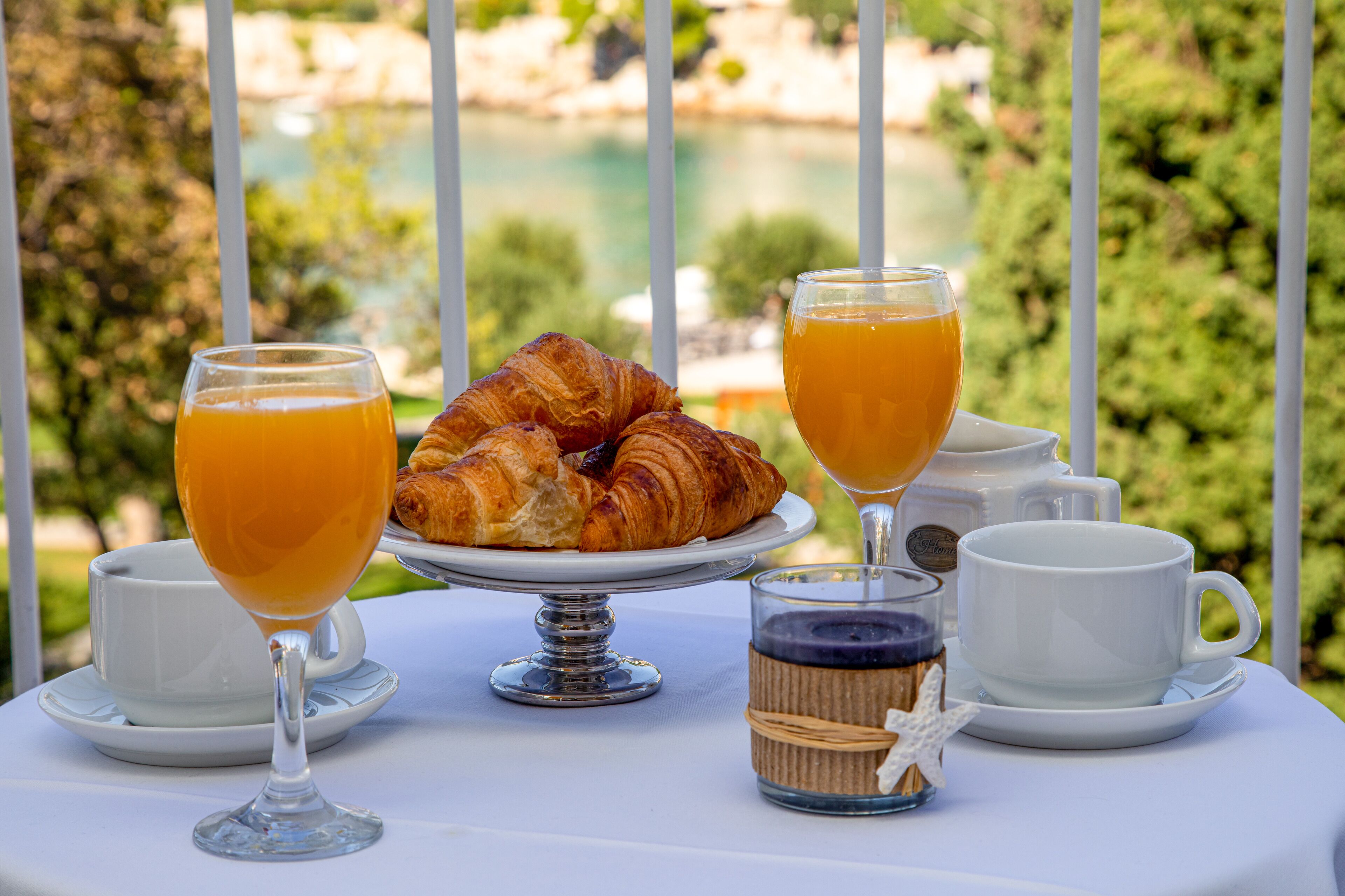 Una mesa con cruasanes, vasos de jugo de naranja y tazas blancas, con una vista panorámica de fondo.