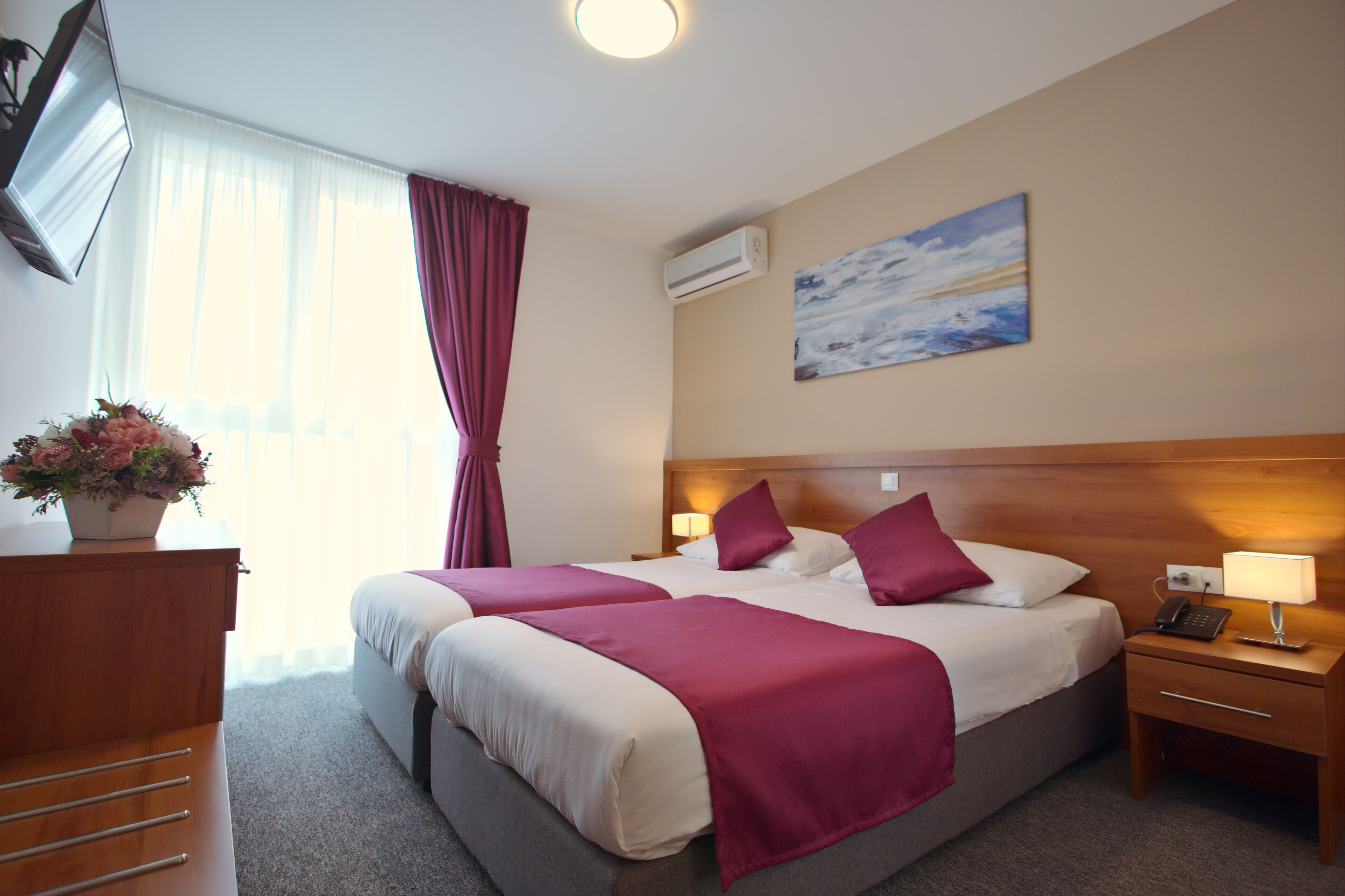 A hotel room with two beds, a wooden headboard, a nightstand with a lamp, a desk with drawers, a potted plant, and a painting on the wall.